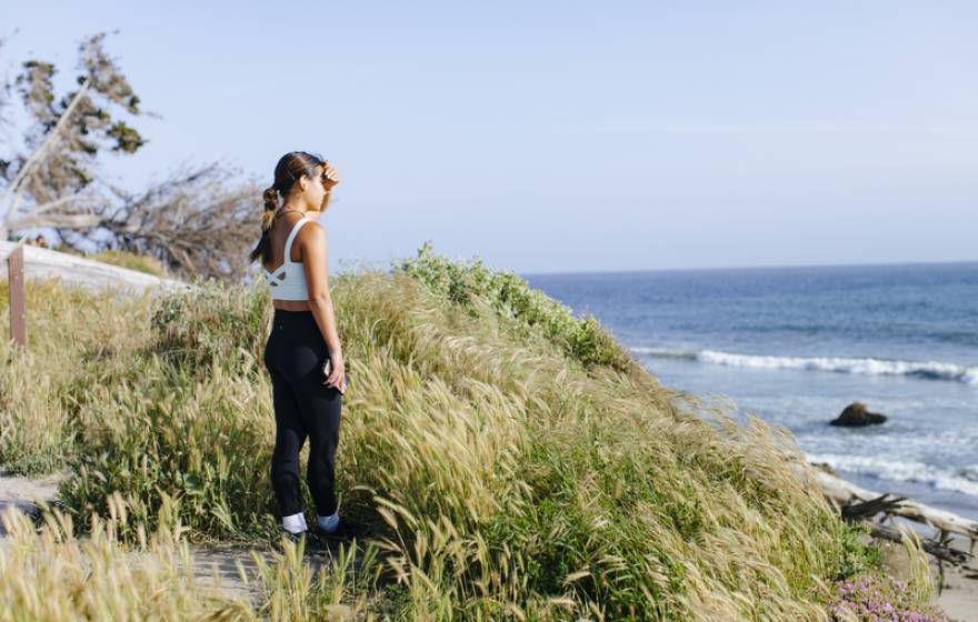 A young woman in exercise attire stands on a grassy bluff overlooking a beach, shading her eyes from the sun