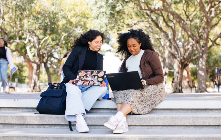 Two students sitting on a stairway outside looking at laptops together and smiling