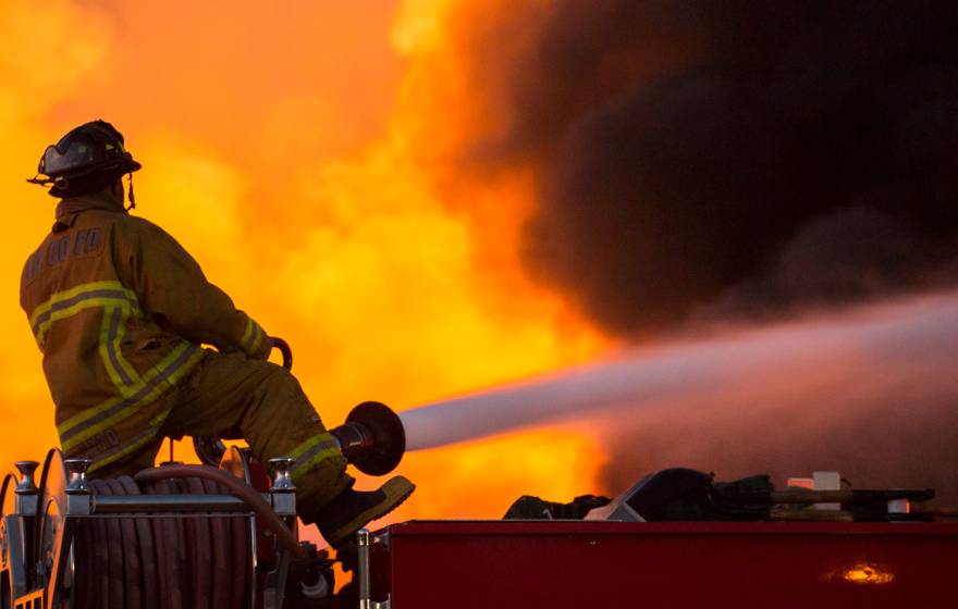 A firefighter straddles a roll of hose, aiming a jet of water into a wall of flames, facing away from the camera