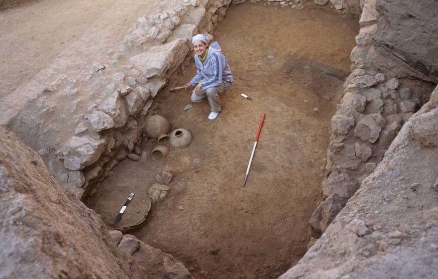 A woman in a very ancient, shallow tomb, looks up from her excavating work, smiling