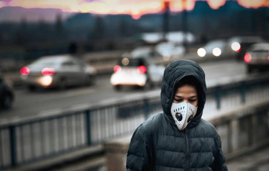 A boy in a hooded jacket standing next to a freeway wearing a respirator mask