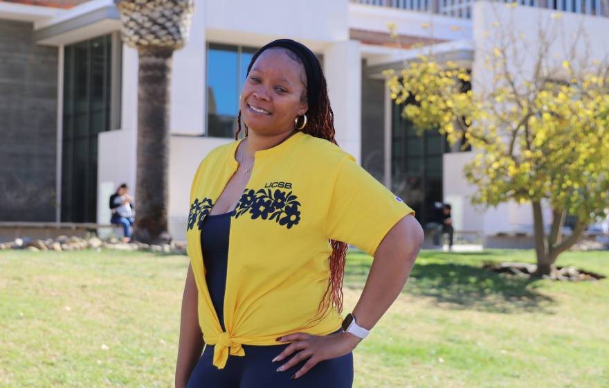 A smiling woman in a yellow UCSB shirt standing in front of a building on campus