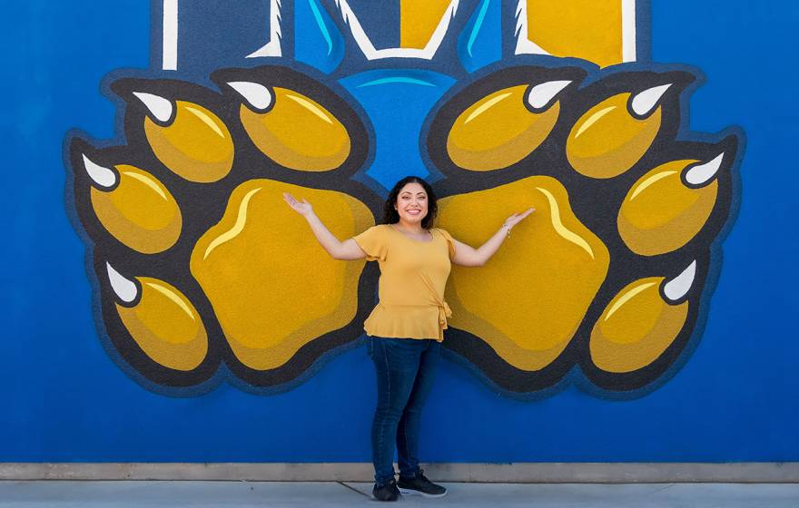 A smiling woman standing in front of a blue wall painted with the UC Merced gold bobcat paws