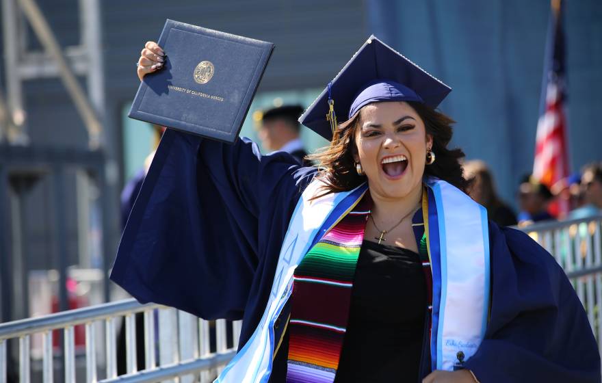 A woman in a cap and gown smiles while holding up her diploma