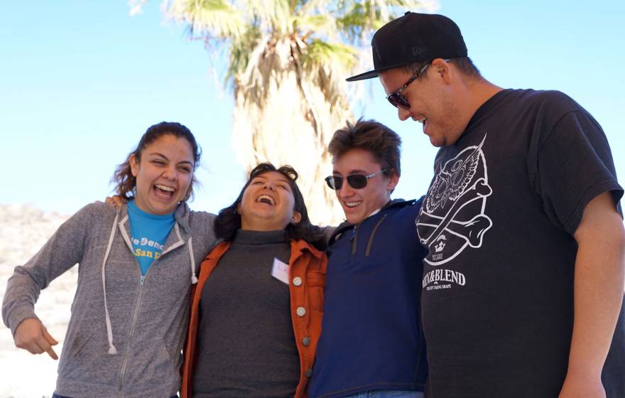 Four students with arms around each other laugh and smile, with a palm tree in the background