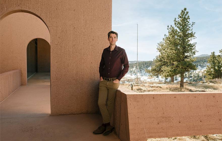 Daniel Swain, wearing khakis and a maroon shirt, leans against a low adobe-colored wall, with a snowy forest in the backdrop and an arched breezeway behind him. 