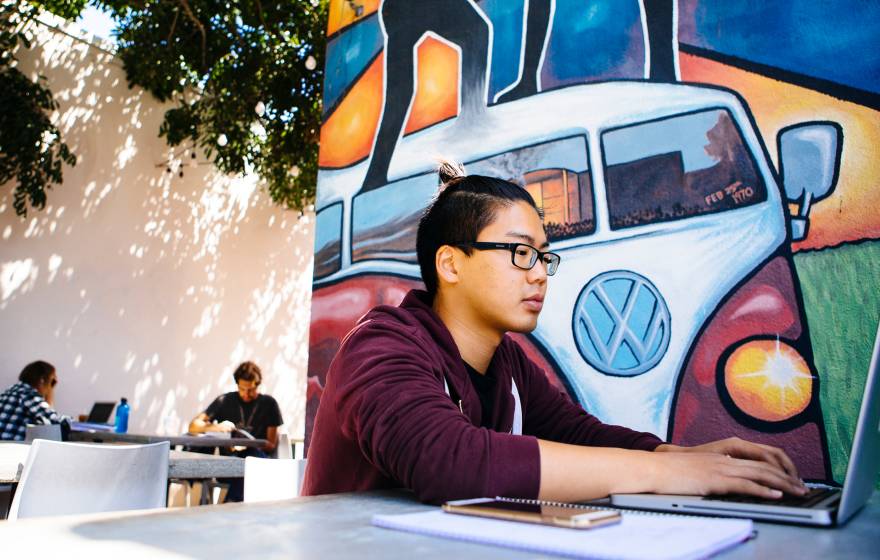 A student working on a laptop at a table outside, in front of a mural showing a Volkswagen van