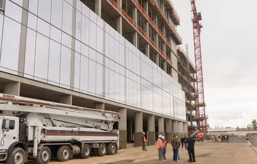 A building  with numerous glass panels under construction on UC Davis' Aggie Square with a construction crew talking below it next to a truck with a crane