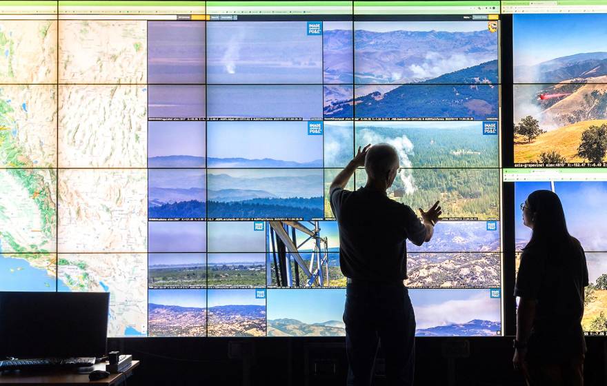 Two people stand in front of a wall of screens showing a grid of footage of natural landscapes, a map of California and some smoke from a wildfire 
