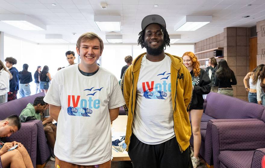 Two UC Santa Barbara students face the camera and smile, wearing t-shirts reading "IV Votes", standing in a busy student lounge.