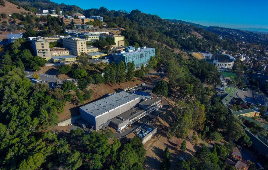 A number of buildings on a tree-covered hillside, including Berkeley Lab, seen from an aerial view on a clear day, Bay in the background