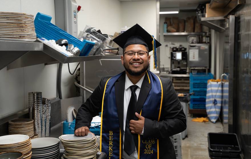 A smiling man in a college graduation cap and gown with a UC Berkeley sash stands in a commercial kitchen in front of a sink and a stack of dishes