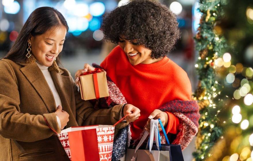 Two women smile and juggled shopping bags next to a Christmas tree.