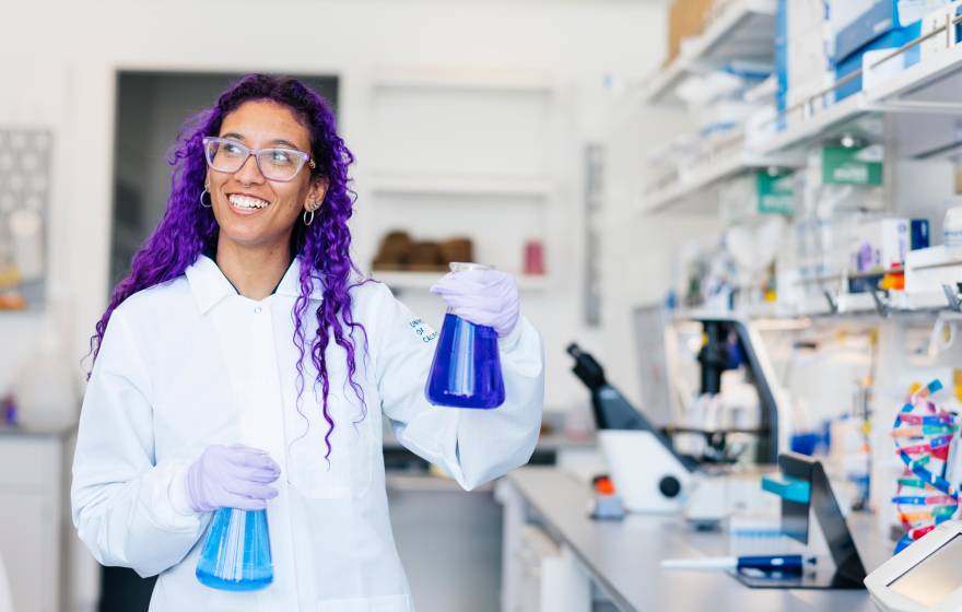 A woman with two flasks of blue chemicals and long purple hair in PPE and a lab coat smiles at a lab table