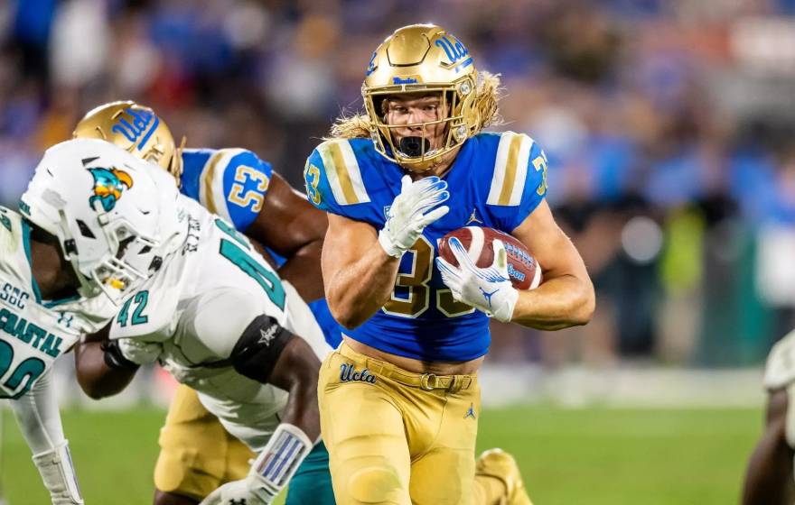 A player in UCLA blue and gold carries the ball and runs by players in white uniforms