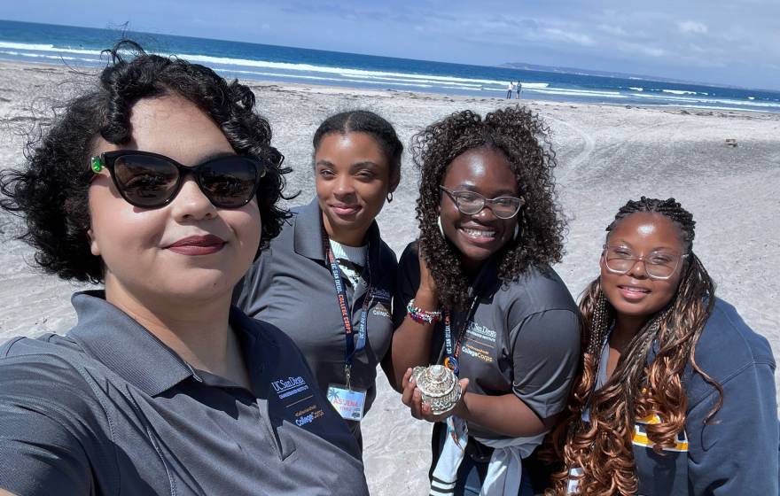 College Corps Four women in gray UC San Diego College Corps polo shirts take a selfie on the beach, smiling