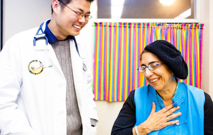 A doctor in a white coat laughs with a patient, who is smiling and holding her hand over her heart.