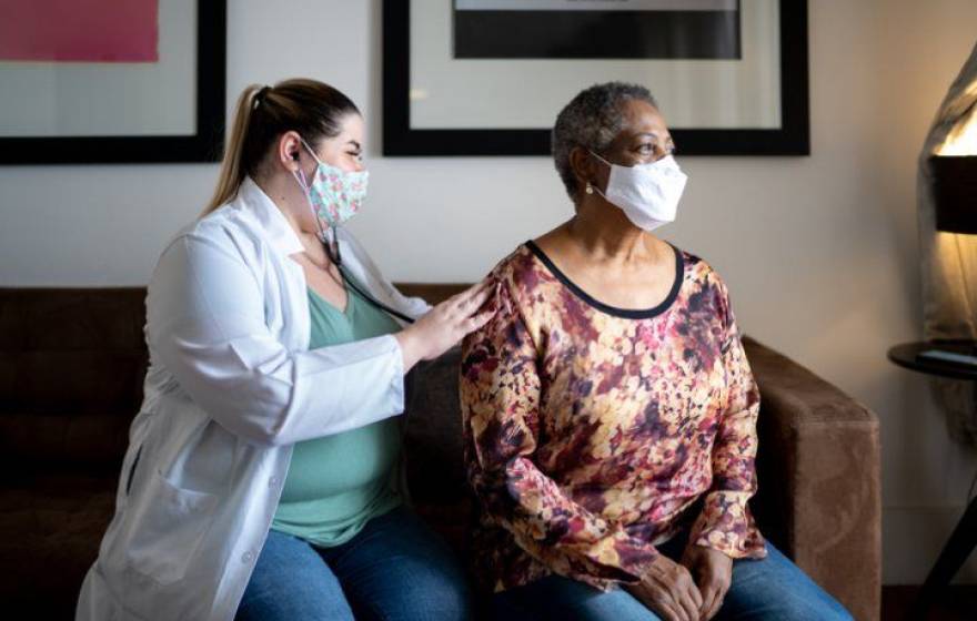 Doctor examining an older Black woman's breathing, both wearing masks