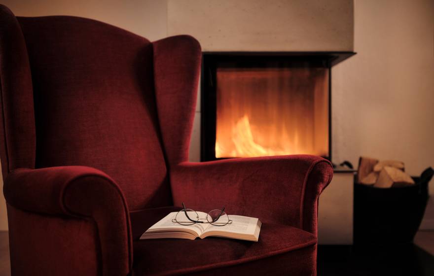 A deep red arm chair with an open book with glasses on top of it next to a fireplace