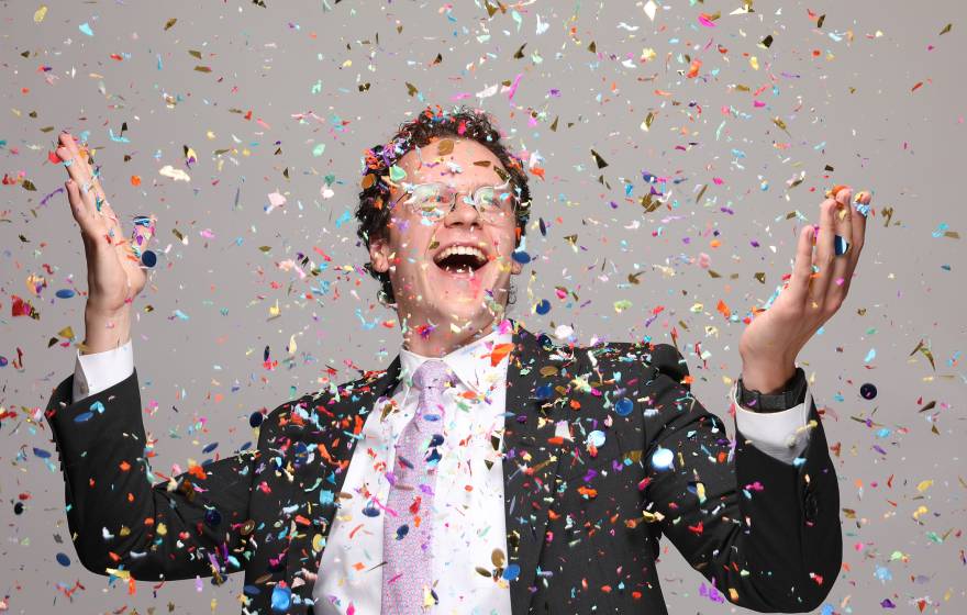 Spelling bee list-maker An ecstatically smiling man with glasses in a suit with a pink tie holds up his arms while confetti falls
