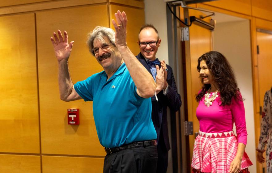 An older man with thick hair and a mustache and glasses throws up his hands as he enters a room and smiles as a woman in a pink dress follows behind, also smilling, and a man behind them smiles, looking on