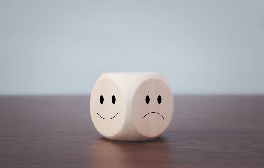 A 6-sided die on a wooden table top. Of the 2 faces visible, one has a happy face and the other has a sad face drawn on it