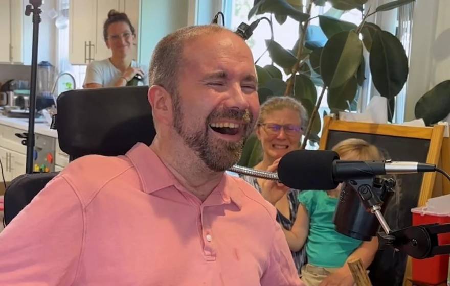 A man with a beard and a pink shirt in his living room grinning, overjoyed in front of a microphone, while his family watches