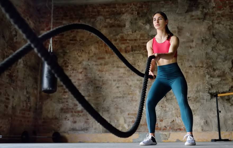 Woman in athletic attire uses battle ropes in a room with an exposed brick wall