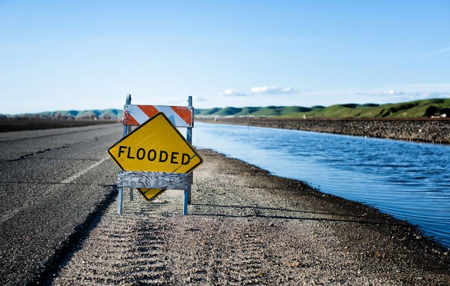 A temporary road sign reads "FLOODED", placed on a highway shoulder next to a canal, with green hills in the background