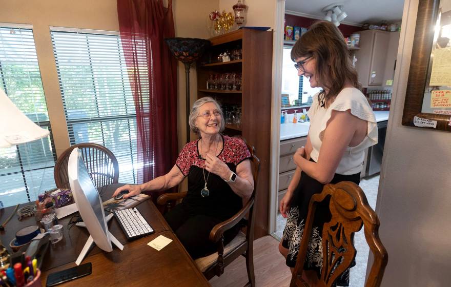 An older woman with gray hair and glasses sits at a computer at the dining room table, smiling and talking with a younger woman standing beside her