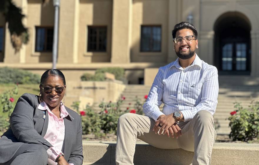 Young Black woman in a gray suit with pink blouse and sunglasses, smiling (Sonya Brooks); and young man (Josiah Beharry) with beard, glasses, khakis, dress shirt, smiling. Both seated on steps at the UC Riverside campus on a bright, sunny day.