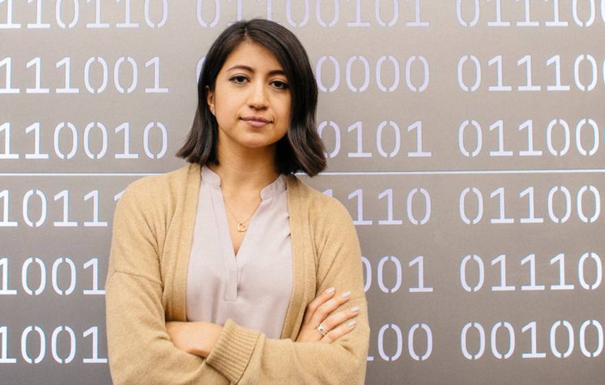 A woman with shoulder length dark hair wearing a cream colored cardigan looks resolutely at the camera with her arms crossed. She's standing in front of a metal panel with sequences of 0s and 1s cut out to simulate computer code. 