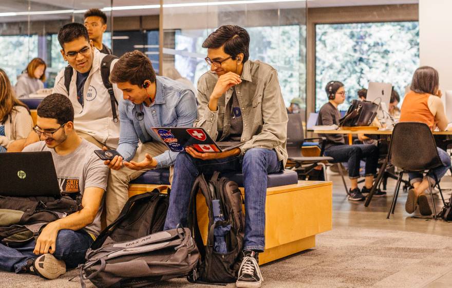 A group of college students work together in a modern campus study area. Some are sitting on benches and the floor with laptops and backpacks, while others collaborate at tables with desktop computers in the background.