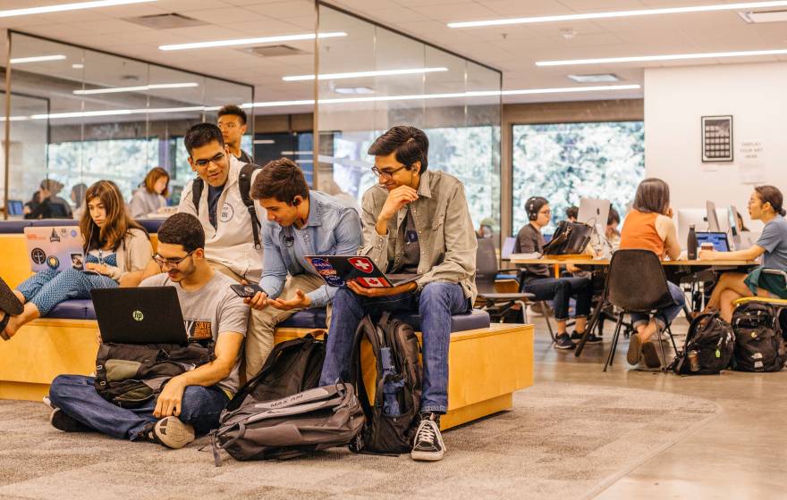 A group of college students work together in a modern campus study area. Some are sitting on benches and the floor with laptops and backpacks, while others collaborate at tables with desktop computers in the background.