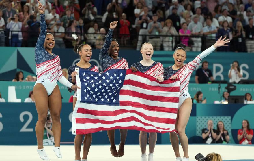 Five gymnasts from Team USA jump and scream while holding the American flag just after winning the 2024 Paris Olympics women's team competition. Credit: James Lang-USA TODAY Sports