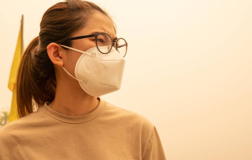 A woman with her hair pulled back wears a N95 mask, looks pensively, yellowish sky around her