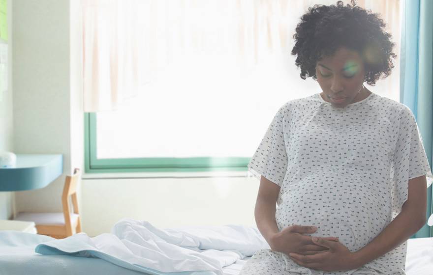 A pregnant woman sits on a hospital bed, wearing a hospital gown, holding her belly from below.