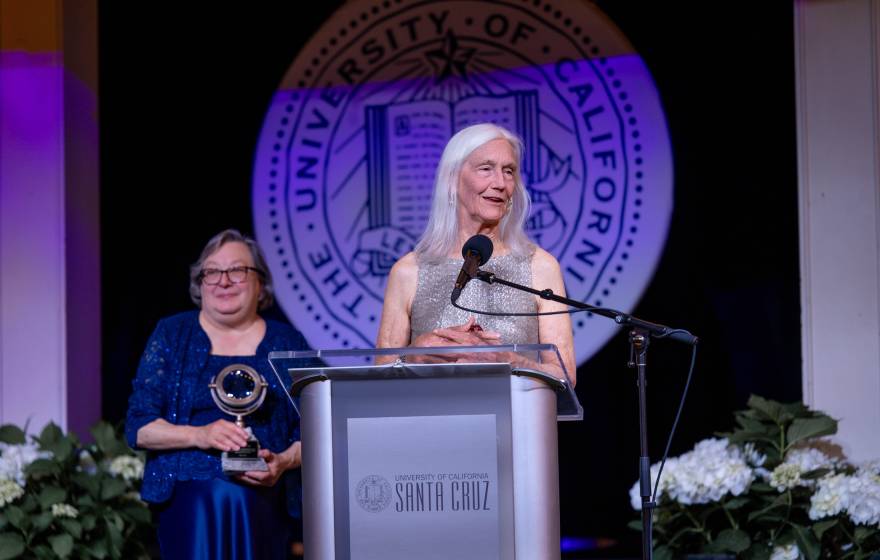 Woman with long white hair (Julie Packard) in silvery dress at a podium; woman in blue dress with short gray hair, Chancellor Larive, holding an award, stands behind