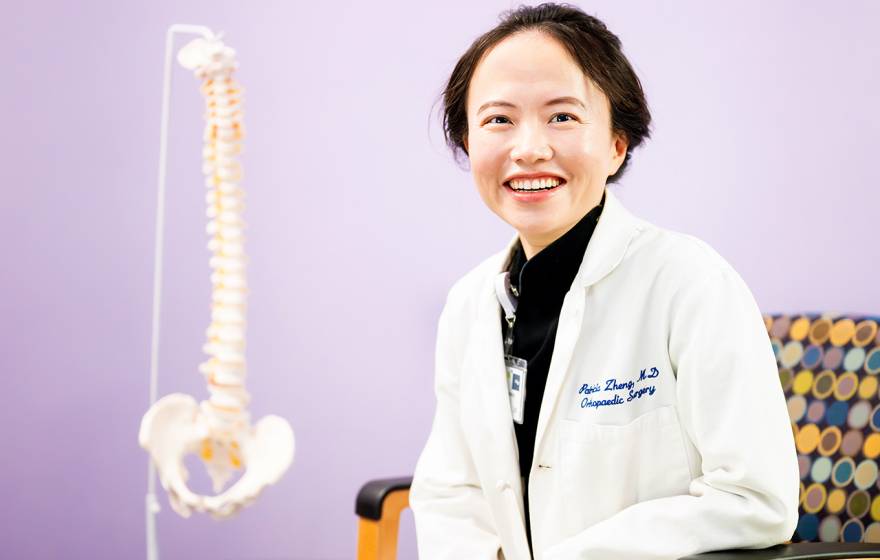 Spine health A woman with short hair in a white doctor's coat smiles in a chair with a model of a human spine behind her against a lilac backdrop