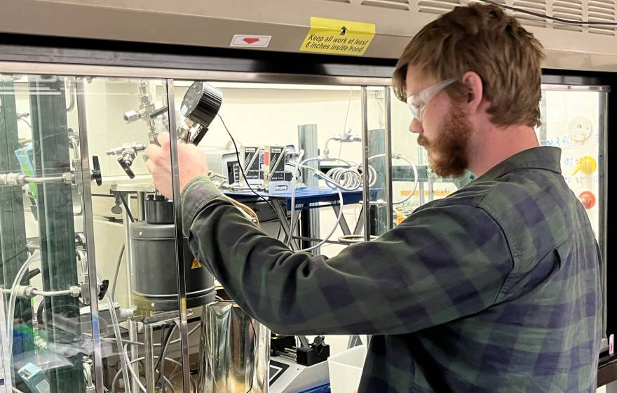 A young bearded man with red hair and protective glasses operates a small machine