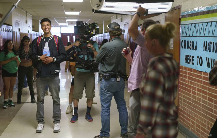 A young man smiling in a busy high school hallway being filmed as part of the movie Rez Ball