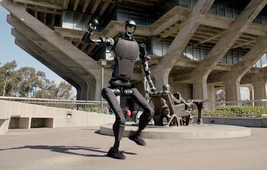 A humanoid robot on the UC San Diego campus in mid-dance move, Geisel Library visible behind