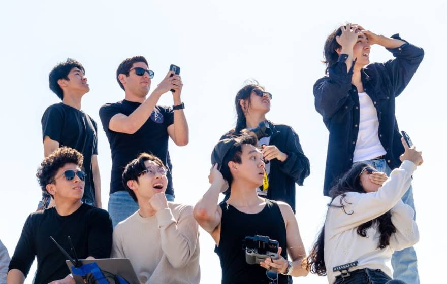 A group of eight people looking at something off camera, four standing, four seated, some gasping, a few with phones recording something, some looking shocked (presumably watching a rocket)