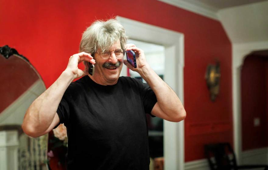 A happy man with white hair and a salt and pepper mustache answers two phones at the same time, one on each ear, laughing