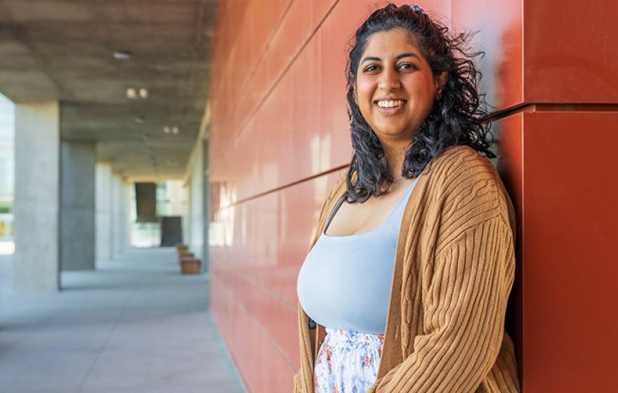 Woman with shoulder-length dark curly hair smiles while standing in an outdoor UC Merced campus corridor