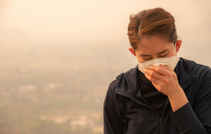 Woman wearing a face mask and coughing into her hand. They sky behind her is hazy with smoke.