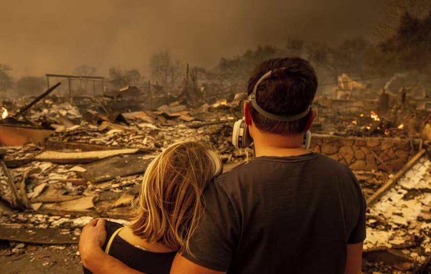 A young man in a mask and woman, photographed from behind, look on at an obliterated house with smoke all around