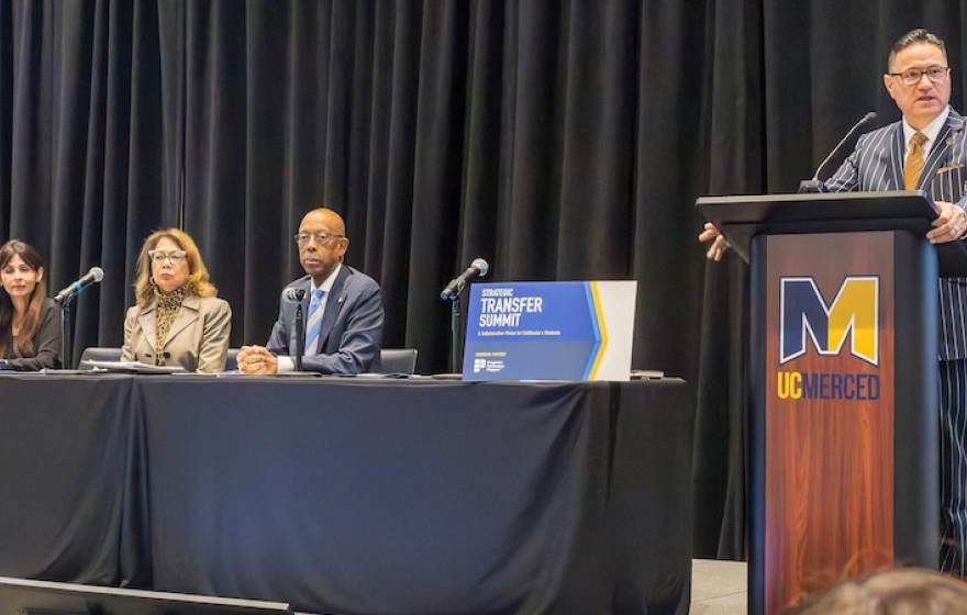 At a table, three people sit in front of microphones, from left: California Community Colleges Chancellor Sonya Christian, California State University Chancellor Mildred Garcia, and UC President Michael Drake; at a lectern, UC Merced Chancellor Juan Sánchez Muñoz stands. Audience is visible a bit in the front of the photo 