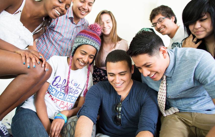 A diverse group of students gather together to look down at something out of frame, like a computer