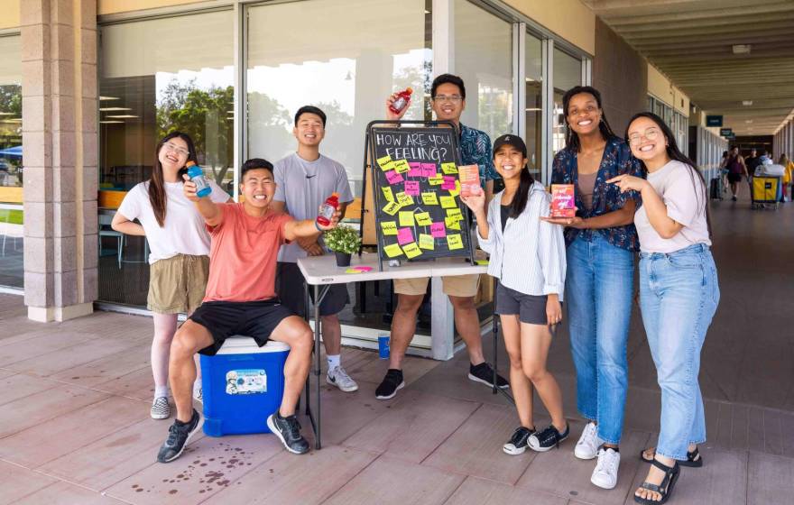 A group of smiling students in front of a dorm with bottles of water and a board that says How Are You Feeling and a number of responses